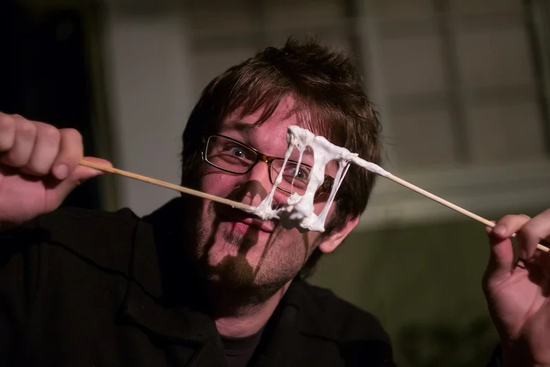 Owen, in glasses, stretching a long strand of toasted marshmallow taffy between two roasting sticks right in front of the camera